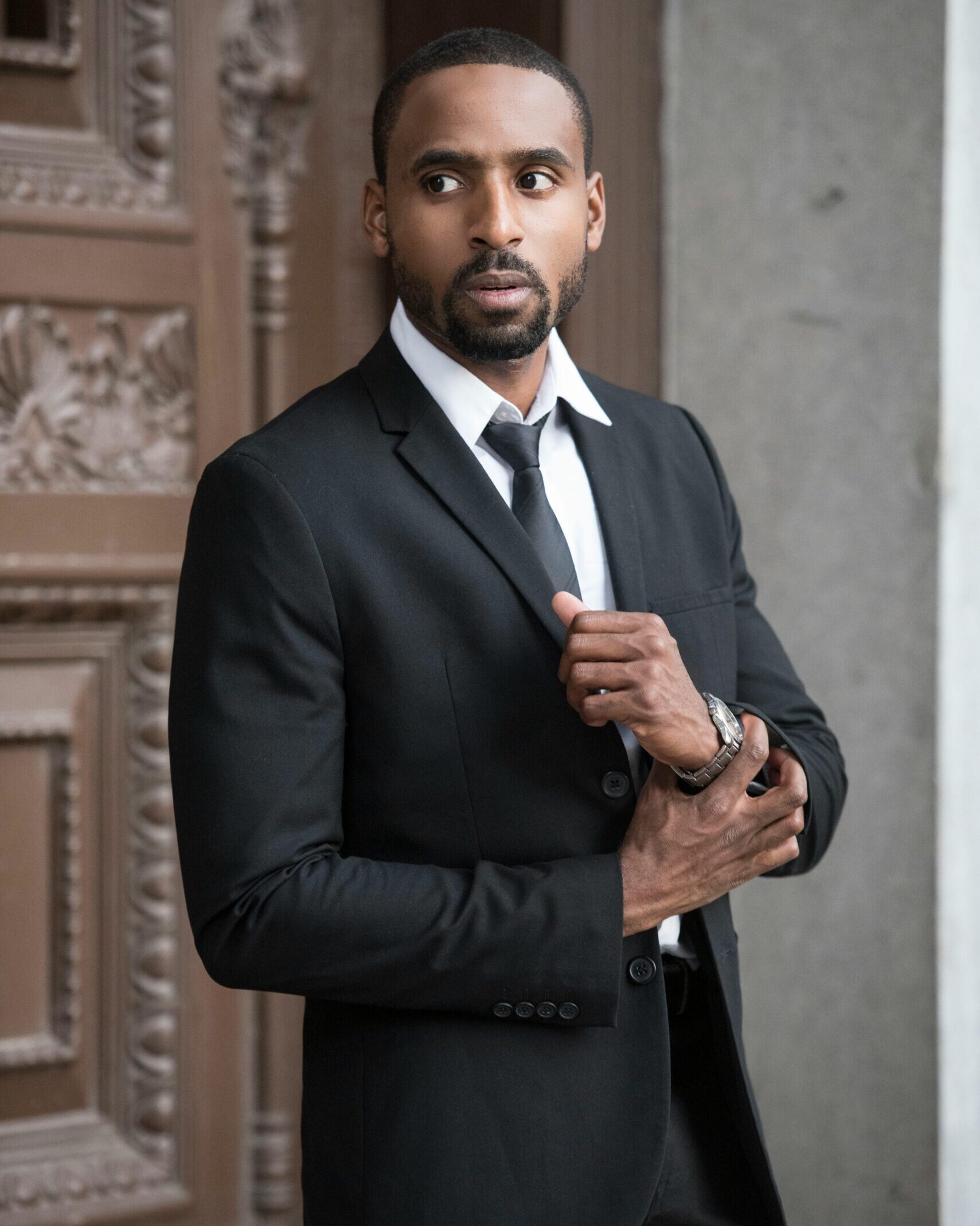 Elegant man in a black suit posing confidently outdoors with a serious expression.